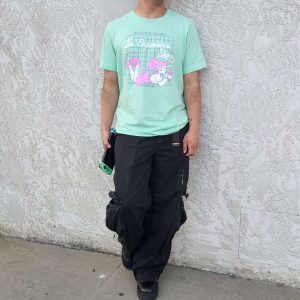 Steven models our merch! He leans against the outer wall of the shop wearing a minty green t-shirt with a white and pink version of our logo.